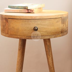 May include: A round wooden nightstand with a single drawer and three legs. The nightstand is made of light brown wood and has a natural finish. The drawer has a metal knob.