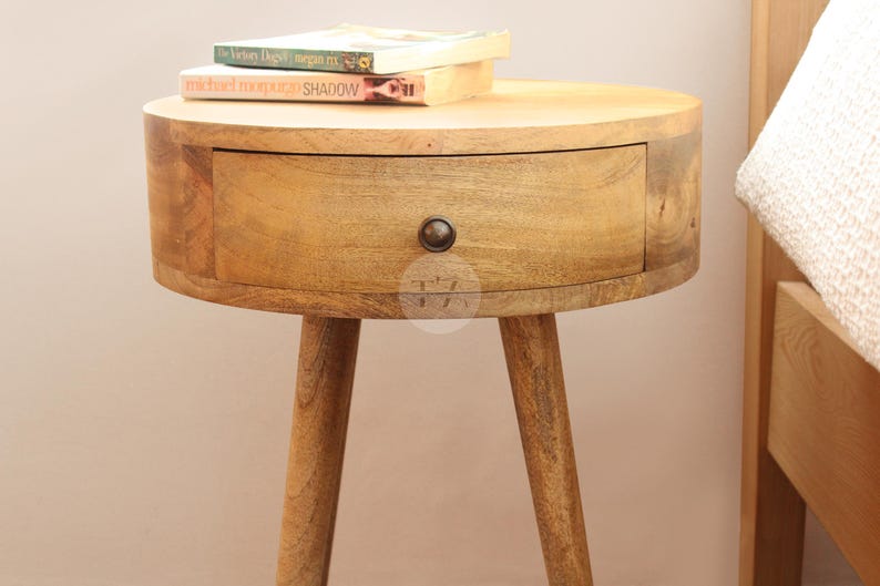 May include: A round wooden nightstand with a single drawer and three legs. The nightstand is made of light brown wood and has a natural finish. The drawer has a metal knob.