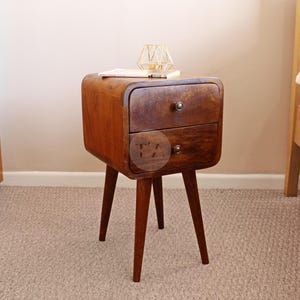May include: A two-drawer wooden nightstand with rounded edges and tapered legs. The nightstand is made of dark wood and has a natural finish.