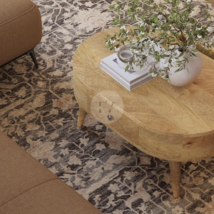 May include: A light wood oval coffee table with a drawer and turned legs. The table is on a patterned rug and has a vase of white flowers on top.