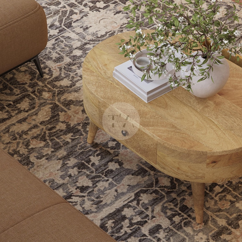 May include: A light wood oval coffee table with a drawer and turned legs. The table is on a patterned rug and has a vase of white flowers on top.