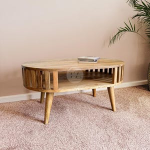 Könnte beinhalten: Ein ovaler Couchtisch aus hellem Holz mit einem unteren Regal und vier Beinen. Der Tisch hat ein Latten-Design an den Seiten und eine glatte Oberfläche.