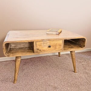 May include: A light wood coffee table with a drawer and open shelves. The table has a curved front edge and tapered legs. A book is resting on the top of the table.