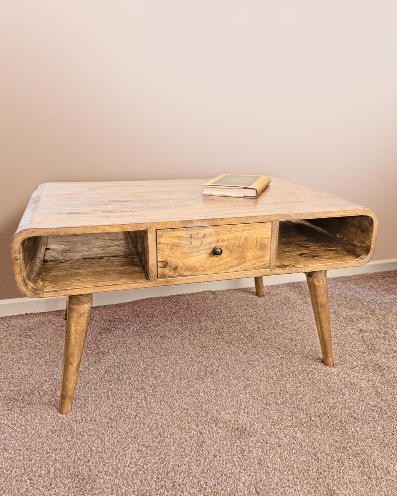 May include: A light wood coffee table with a drawer and open shelves. The table has a curved front edge and tapered legs. A book is resting on the top of the table.