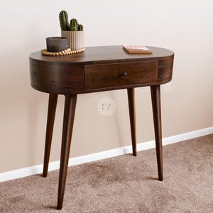 May include: A dark brown, oval-shaped side table with a single drawer and four tapered legs. The table is made of wood and has a smooth finish. A small cactus, a candle, and a book are on top.