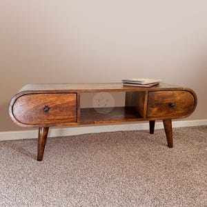 Puede incluir: Una mesa de centro de estilo moderno de mediados de siglo con una parte superior redondeada de forma ovalada. La mesa está hecha de madera oscura y tiene dos cajones y un estante. La mesa está sobre una alfombra de color marrón claro.