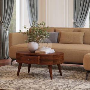 May include: A brown oval coffee table with a single drawer and four legs. The table is in a living room setting with a beige sofa and a vase of white flowers.