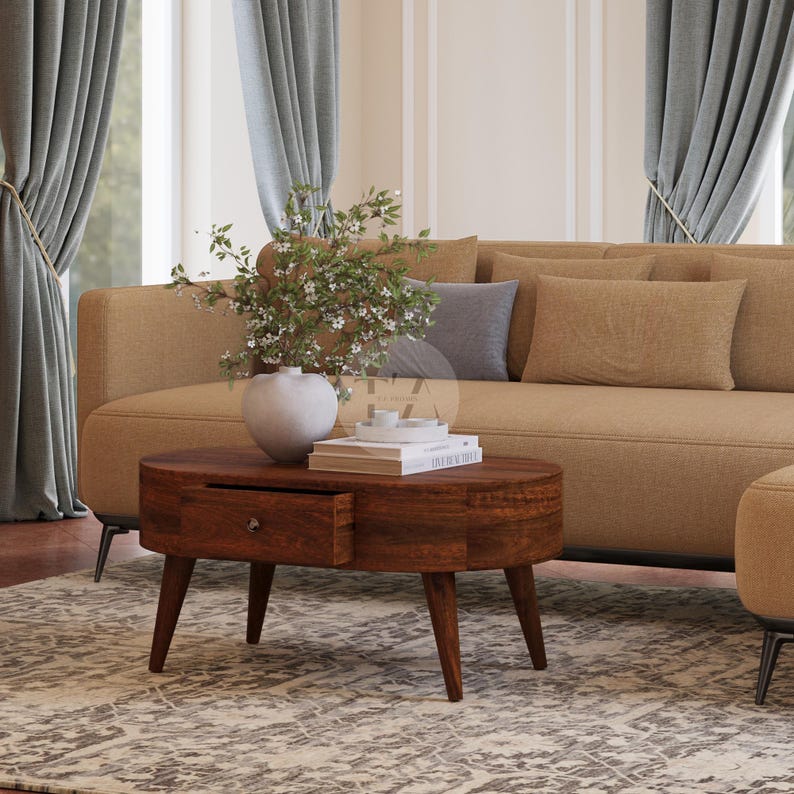 May include: A brown oval coffee table with a single drawer and four legs. The table is in a living room setting with a beige sofa and a vase of white flowers.