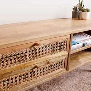 Pu&ograve; includere: Una consolle multimediale in legno chiaro con due cassetti con frontali in canna intrecciata e tiretti in ottone. La consolle ha uno scaffale aperto con due libri sopra.