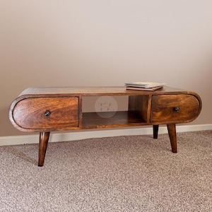 May include: A mid-century modern style coffee table with a rounded, oval-shaped top. The table has two drawers and a shelf, and is made of dark brown wood. The table is sitting on a light brown carpet.