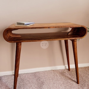 May include: A wooden desk with a curved design and a lower shelf. The desk has four tapered legs and a natural wood finish. A small stack of books rests on the top surface. The desk is approximately 48 inches long.