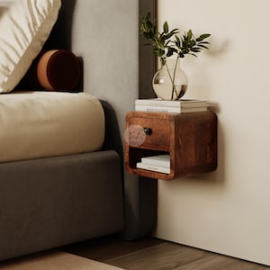Può includere: Un comodino in legno sospeso con un cassetto e un ripiano aperto. Il comodino è di colore marrone caldo e ha bordi arrotondati. Un piccolo vaso di vetro con foglie verdi è posto sopra, insieme a una pila di libri. Il comodino è montato su una parete bianca.