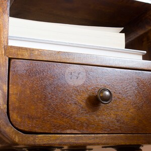 May include: A wooden nightstand with a curved top shelf and a drawer with a metal knob. The shelf has a stack of books on it.