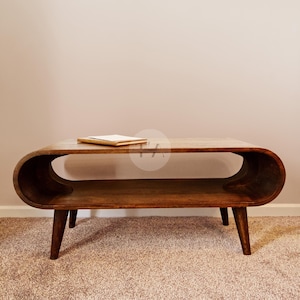 Op de afbeelding: Een donkerbruine houten salontafel met een gebogen bovenblad en een lager plankje. De tafel heeft vier poten en staat op een beigekleurig tapijt.