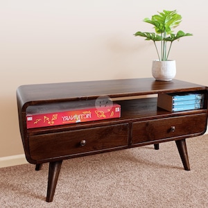 May include: A dark brown wooden TV stand with two drawers and a shelf. The stand has rounded edges and tapered legs. A red game box and a stack of blue books are on the shelf. A potted plant sits on top.