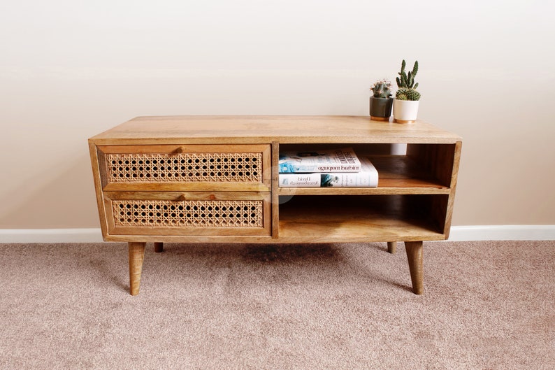 Pu&ograve; includere: Un mobile TV in legno chiaro con due cassetti con frontali in vimini intrecciato. Il mobile ha due ripiani aperti e poggia su quattro gambe affusolate. Due libri sono sul ripiano superiore.