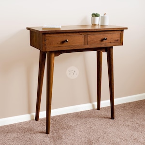 May include: A wooden console table with two drawers and tapered legs. The table is a warm brown colour and has two small black knobs on the drawers. A small potted plant and a white container sit on top. The table is approximately 76 cm tall.