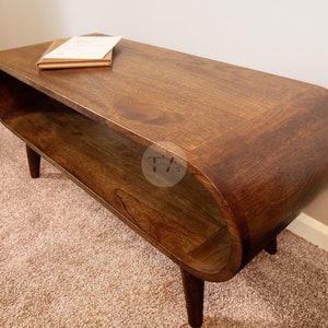 Op de afbeelding: Een donkerbruine houten salontafel met een gebogen, ovaalvormig blad en een lager plankje. De tafel heeft een mid-century modern design en staat op een beigekleurig tapijt.