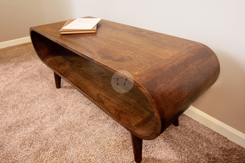 Op de afbeelding: Een donkerbruine houten salontafel met een gebogen, ovaalvormig blad en een lager plankje. De tafel heeft een mid-century modern design en staat op een beigekleurig tapijt.