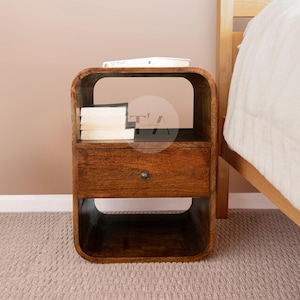 May include: A rectangular wooden nightstand with rounded corners, featuring a single central drawer with a small circular knob, an open storage compartment above the drawer containing a stack of books, and an open storage space below the drawer.