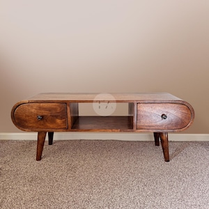 Op de afbeelding: Een lage houten salontafel met een rijke bruine afwerking, voorzien van afgeronde uiteinden, twee lades met kleine donkere knoppen en een open opbergplank in het midden. De tafel rust op vier taps toelopende houten poten en heeft een glad oppervlak.