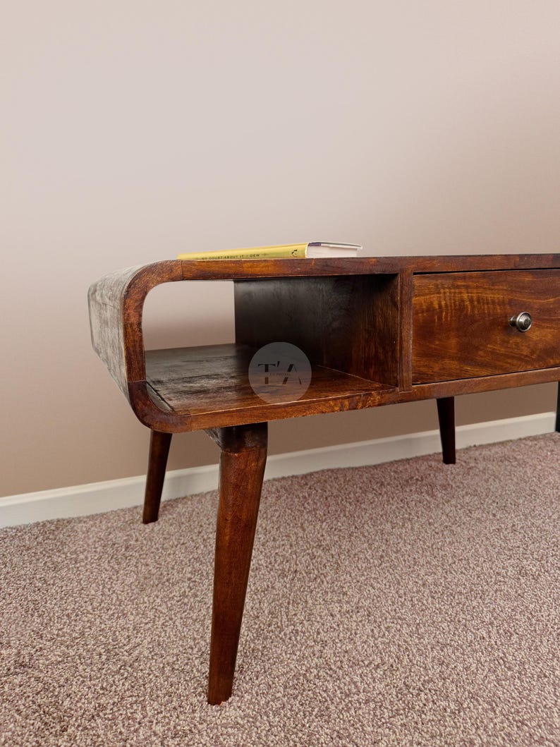 May include: A dark brown wooden coffee table with a curved shelf and a drawer. The table has four legs and is sitting on a light brown carpet.