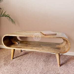 Könnte beinhalten: Ein hellbrauner Couchtisch aus Holz mit einem glatten, durchgehend geschwungenen Rahmen, der eine offene Ablagefläche bildet. Der Tisch hat vier konische Beine und eine rechteckige Oberfläche, auf der ein kleines geschlossenes Buch liegt.