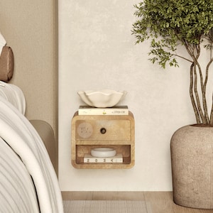 May include: A wall-mounted light wood bedside table with a single drawer and an open storage shelf below. The top surface holds a cream-coloured scalloped bowl and a book. A second book sits on the lower shelf. The unit has a rounded square shape.