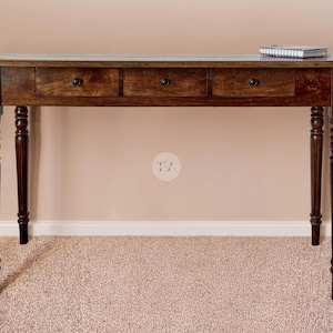 May include: A dark brown wooden desk with three drawers and turned legs. A small notebook rests on the desk's surface. The desk is set against a neutral-coloured wall and carpeted floor.