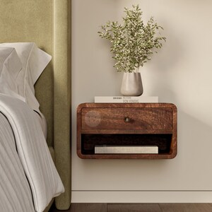 K&ouml;nnte beinhalten: Ein dunkelbraunes, schwebendes Nachttischchen aus Holz mit einer Schublade und einem Regal. Der Nachttisch ist an einer wei&szlig;en Wand neben einem Bett mit einem gr&uuml;nen Kopfteil montiert. Ein Buch mit dem Titel "DREISER,  SELECTED  VON  VENEDIG" liegt auf dem Nachttisch. Eine Vase mit wei&szlig;en Blumen steht auf dem Regal.