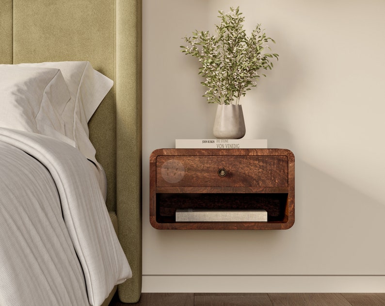K&ouml;nnte beinhalten: Ein dunkelbraunes, schwebendes Nachttischchen aus Holz mit einer Schublade und einem Regal. Der Nachttisch ist an einer wei&szlig;en Wand neben einem Bett mit einem gr&uuml;nen Kopfteil montiert. Ein Buch mit dem Titel "DREISER,  SELECTED  VON  VENEDIG" liegt auf dem Nachttisch. Eine Vase mit wei&szlig;en Blumen steht auf dem Regal.