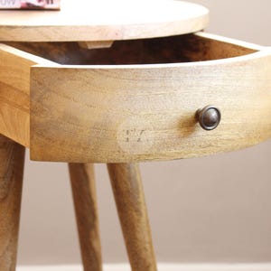 May include: A round wooden side table with a single drawer and turned legs. The drawer has a dark brown knob and is open, revealing the interior.