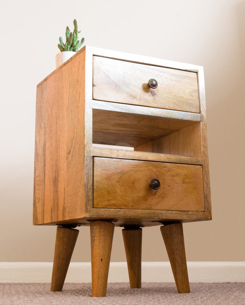 K&ouml;nnte beinhalten: Ein Nachttisch aus Holz mit zwei Schubladen und einem Regal. Der Nachttisch hat eine hellbraune Oberfl&auml;che und vier konische Beine.