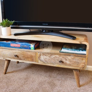 May include: A light wood TV stand with two drawers and a shelf. The stand has a curved front and rounded legs. A flat screen TV is sitting on top of the stand. A board game and a book are on the shelf.