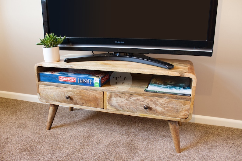 May include: A light wood TV stand with two drawers and a shelf. The stand has a curved front and rounded legs. A flat screen TV is sitting on top of the stand. A board game and a book are on the shelf.