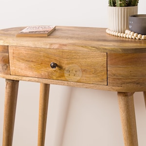 May include: Oval wooden side table with a single drawer and four tapered legs. The table is made of light-colored wood with visible grain. A small book, a potted plant, and a candle sit on top.
