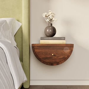 Può includere: Un comodino sospeso semicircolare montato a parete, realizzato in legno marrone scuro con un cassetto dotato di un piccolo pomello dorato. La superficie ospita due libri impilati e un vaso grigio rotondo con fiori bianchi essiccati.