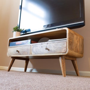 May include: A light wood TV stand with two drawers and a shelf. The stand has a curved front and four legs. The stand is sitting on a beige carpet. There are two game boxes on the shelf, one says "MONOPOLY" and the other says "Pictionary".