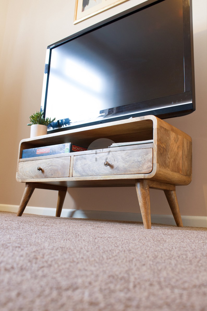 May include: A light wood TV stand with two drawers and a shelf. The stand has a curved front and four legs. The stand is sitting on a beige carpet. There are two game boxes on the shelf, one says "MONOPOLY" and the other says "Pictionary".
