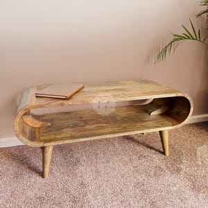 May include: A wooden coffee table with a unique, curved design. The table has a natural wood finish and a lower shelf for storage. A closed book rests on the tabletop. The table is supported by four tapered legs.