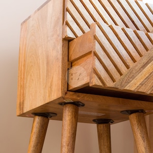 May include: A wooden side table with a drawer and four legs. The drawer has a geometric pattern carved into the front. The table is made of light brown wood and has a natural finish.