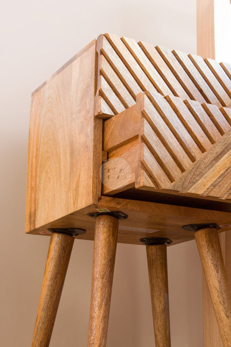 May include: A wooden side table with a drawer and four legs. The drawer has a geometric pattern carved into the front. The table is made of light brown wood and has a natural finish.
