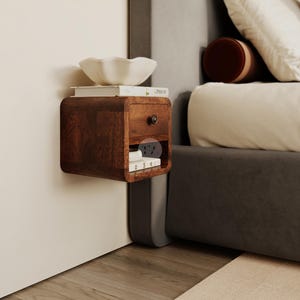 May include: A dark brown, wall-mounted nightstand with a drawer and a small shelf. A white bowl and books sit on top. The nightstand is next to a bed with pillows and a rug.
