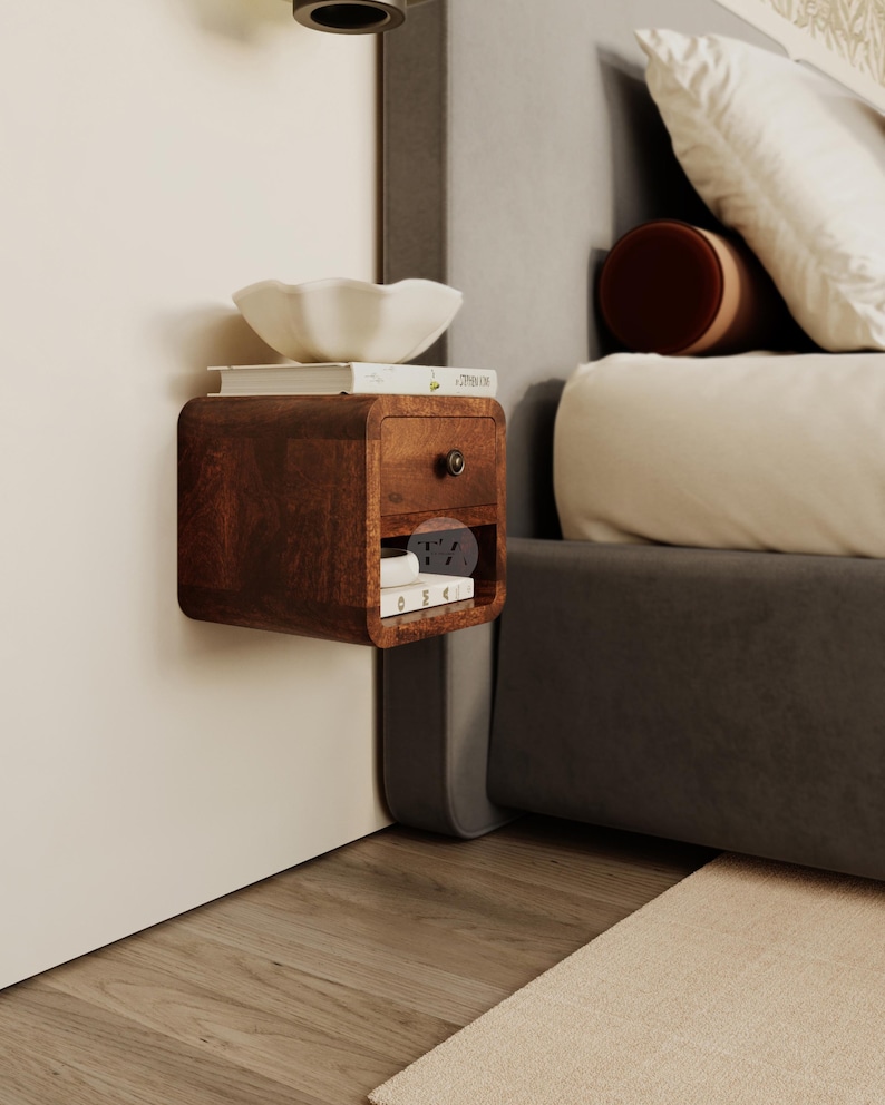 May include: A dark brown, wall-mounted nightstand with a drawer and a small shelf. A white bowl and books sit on top. The nightstand is next to a bed with pillows and a rug.