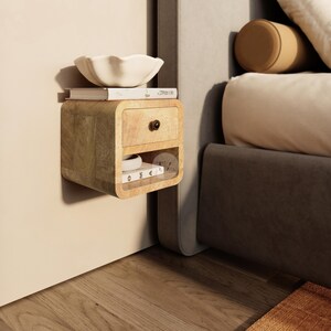 May include: A floating wooden nightstand with a drawer and open shelf. A white bowl and books sit on top. The nightstand is mounted on a beige wall next to a bed with a gray base and white bedding. A brown bolster pillow rests on the bed.
