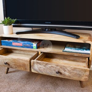 May include: A wooden TV stand with two drawers and a shelf. The drawers are open and the shelf is holding a board game and a book. The TV stand is on a carpet.