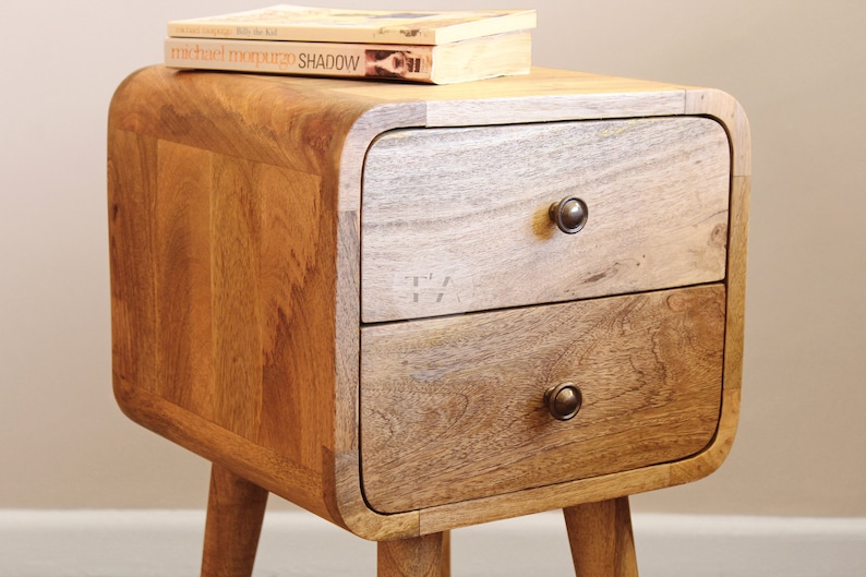 May include: A wooden bedside table with two drawers and rounded edges. The bedside table has a light brown finish and features two black metal knobs. The bedside table is sitting on a light brown floor.