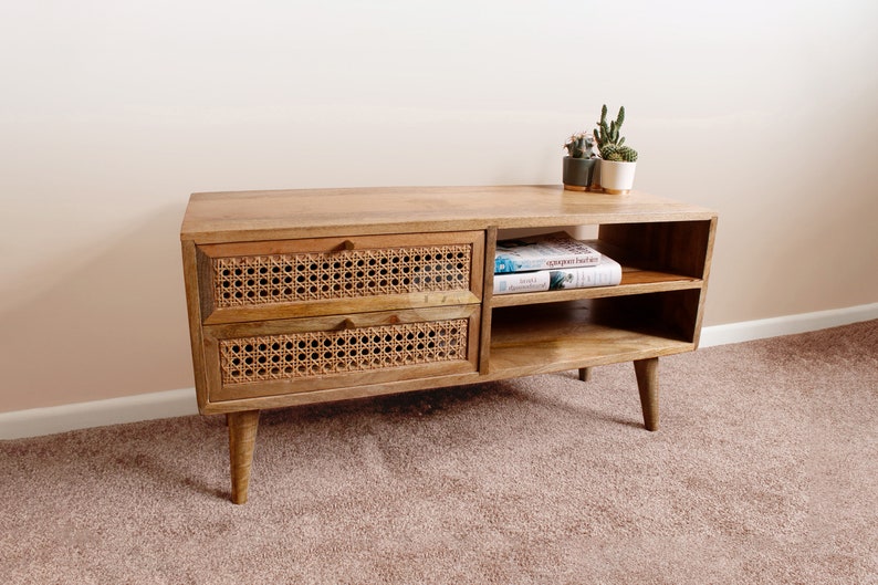 Pu&ograve; includere: Un mobile TV in legno chiaro con due cassetti con frontali in vimini intrecciato e due ripiani aperti. Il mobile ha un design moderno con gambe affusolate ed &egrave; posizionato su un tappeto beige.