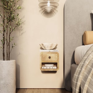May include: A wall-mounted wooden nightstand with a drawer and open shelf, holding a small white object and a book. A decorative bowl sits on top. A potted plant and a layered pendant light are also visible.