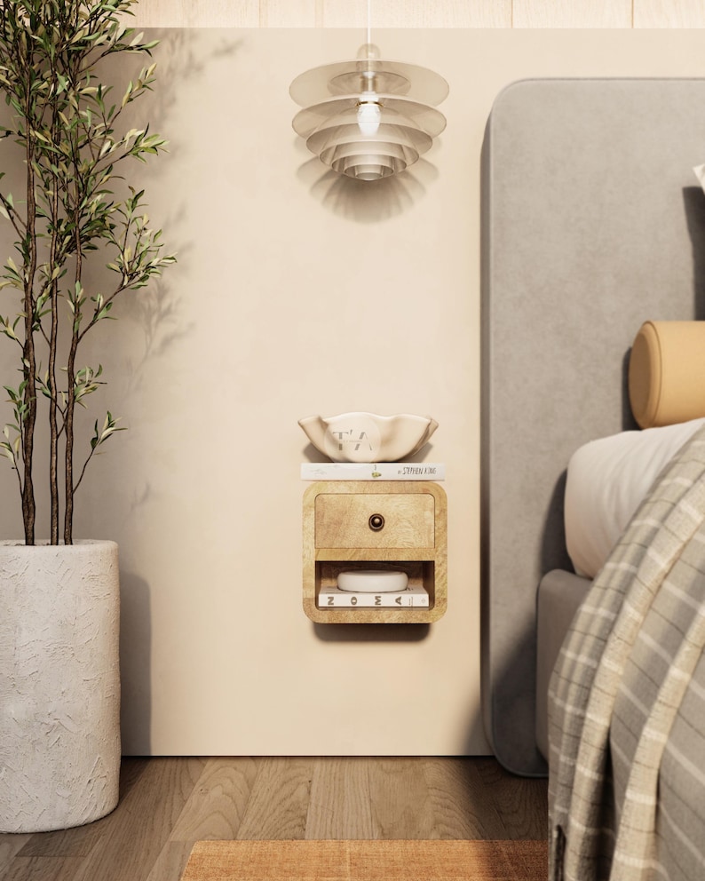 May include: A wall-mounted wooden nightstand with a drawer and open shelf, holding a small white object and a book. A decorative bowl sits on top. A potted plant and a layered pendant light are also visible.